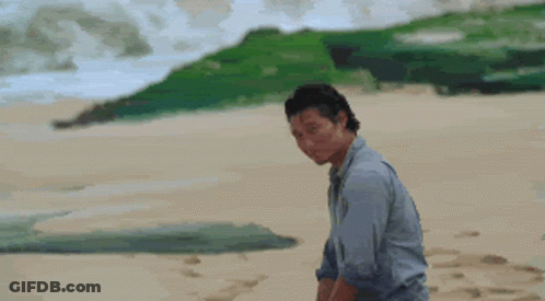 A Man In A Blue Shirt Is Standing On A Sandy Beach Looking At Something GIF