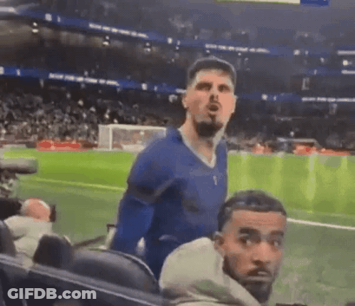 A Man In A Blue Shirt Is Standing On A Soccer Field While Another Man Looks On . GIF