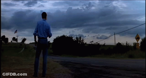 A Man In A Blue Shirt Is Standing On The Side Of A Road In Front Of A Road Sign GIF