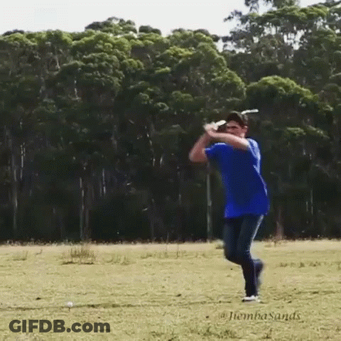 A Man In A Blue Shirt Is Swinging A Golf Club In A Field With Trees In The Background GIF