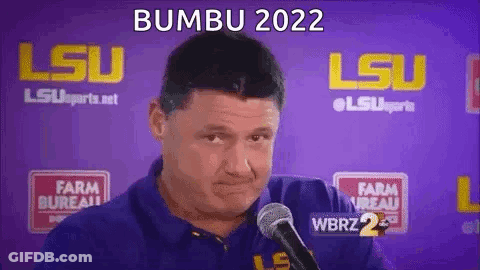 A Man In A Blue Shirt Is Talking Into A Microphone In Front Of A Purple Background . GIF