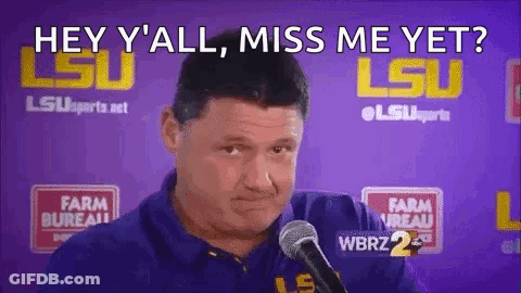 A Man In A Blue Shirt Is Talking Into A Microphone In Front Of A Purple Wall . GIF