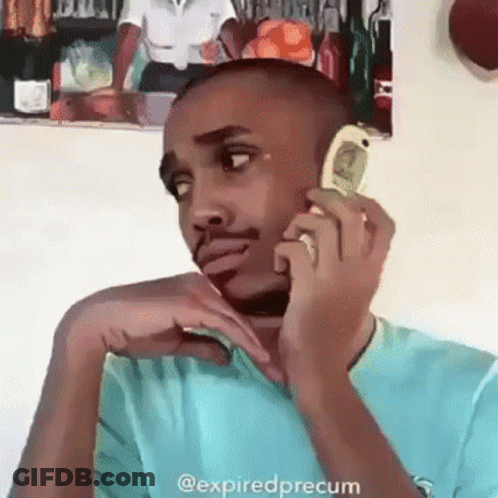 A Man In A Blue Shirt Is Talking On A Cell Phone With A Picture Of A Woman On The Wall Behind Him . GIF
