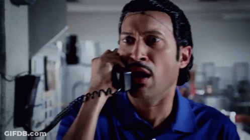 A Man In A Blue Shirt Is Talking On A Phone With A Surprised Look On His Face GIF