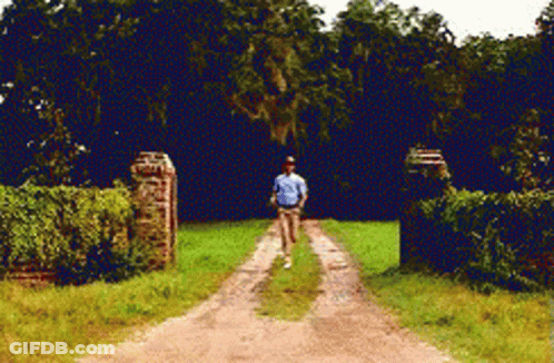 A Man In A Blue Shirt Is Walking Down A Dirt Road Surrounded By Trees GIF