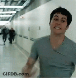 A Man In A Blue Shirt Is Walking Down A Hallway With People Walking In The Background . GIF