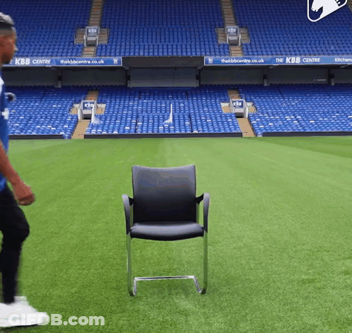 A Man In A Blue Shirt Is Walking On A Soccer Field With A Chair In The Foreground That Says Kbb Centre GIF