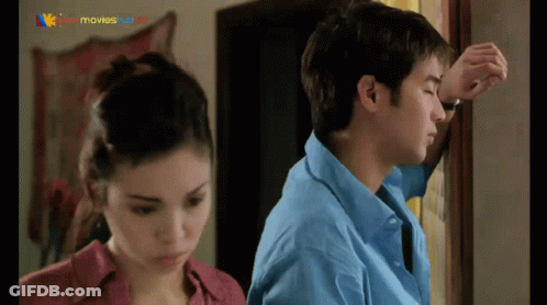 A Man In A Blue Shirt Leans Against A Wall While A Woman In A Red Shirt Looks On GIF