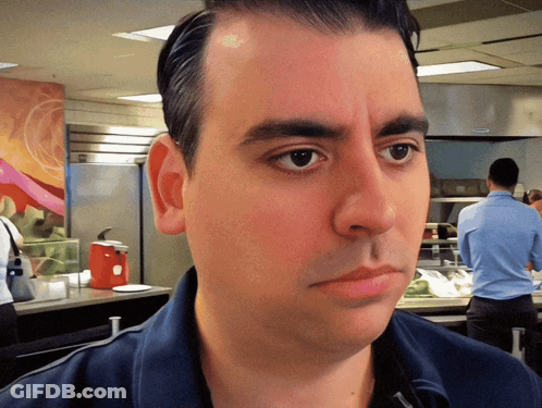 A Man In A Blue Shirt Looks At The Camera With A Red Ketchup Dispenser In The Background GIF
