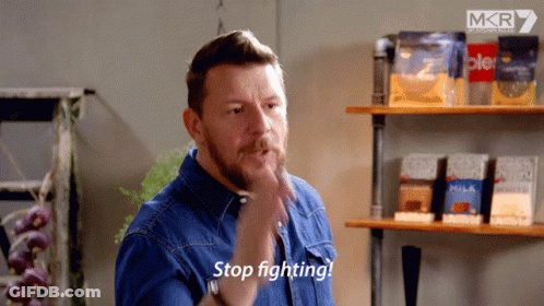 A Man In A Blue Shirt Says Stop Fighting In Front Of Some Shelves GIF