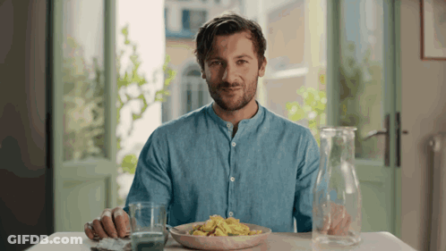 A Man In A Blue Shirt Sits At A Table With A Plate Of Food GIF