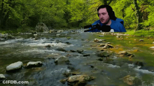 A Man In A Blue Shirt Sits In Front Of A Stream GIF