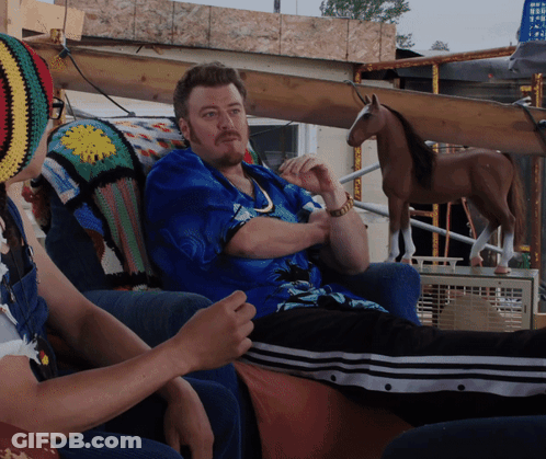A Man In A Blue Shirt Sits On A Couch With A Woman In A Crocheted Hat GIF
