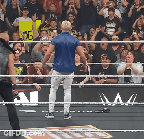 A Man In A Blue Shirt Stands In A Wrestling Ring With A Crowd Behind Him GIF