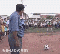 A Man In A Blue Shirt Stands In Front Of A Soccer Ball On A Dirt Field GIF