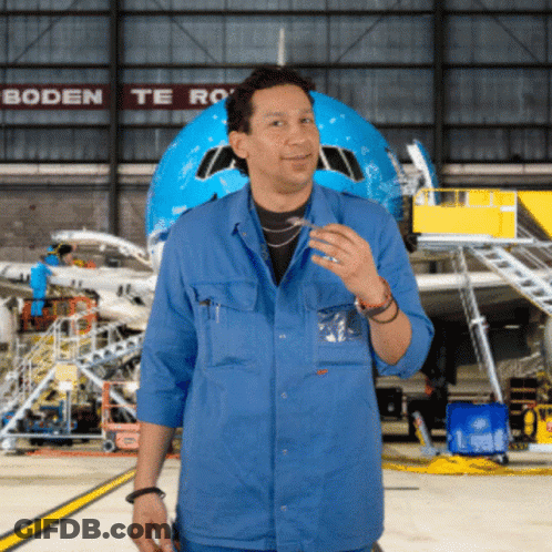 A Man In A Blue Shirt Stands In Front Of An Airplane In A Hangar With Boden Te Ro Written On The Wall Behind Him GIF