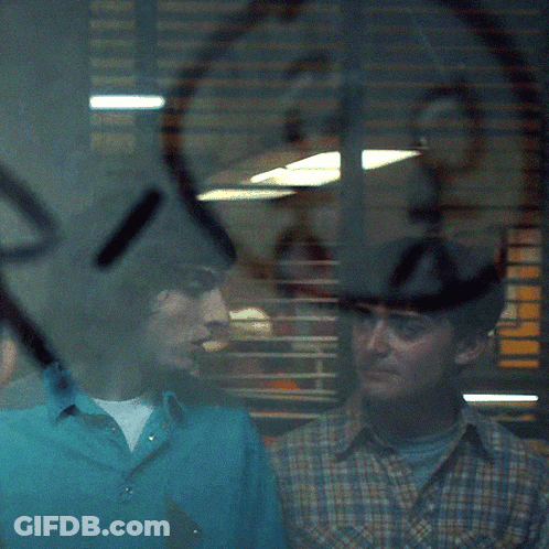 A Man In A Blue Shirt Stands Next To Another Man In A Plaid Shirt In Front Of A Window GIF
