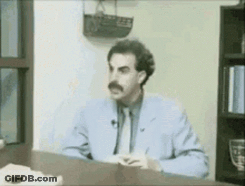 A Man In A Blue Suit And Tie Is Sitting At A Desk In Front Of A Window . GIF