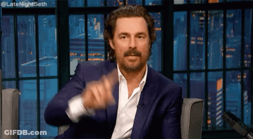 A Man In A Blue Suit And White Shirt Is Sitting In A Chair And Pointing At The Camera . GIF