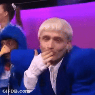 A Man In A Blue Suit Is Covering His Mouth With His Hand While Sitting In Front Of A Purple Wall . GIF