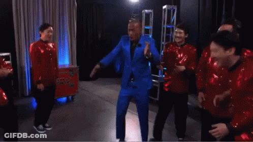 A Man In A Blue Suit Is Dancing With A Group Of Men In Red Sequins GIF