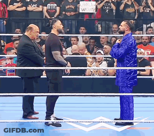 A Man In A Blue Suit Stands In The Middle Of A Wrestling Ring With Two Other Men GIF