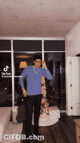 A Man In A Blue Sweater And Black Pants Is Standing In Front Of A Christmas Tree In A Living Room GIF