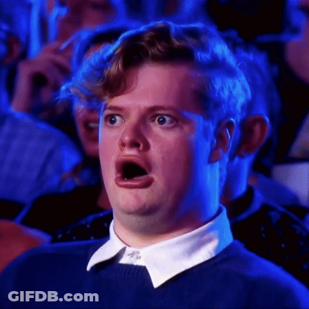 A Man In A Blue Sweater And White Collar Is Making A Surprised Face While Sitting In A Crowd . GIF