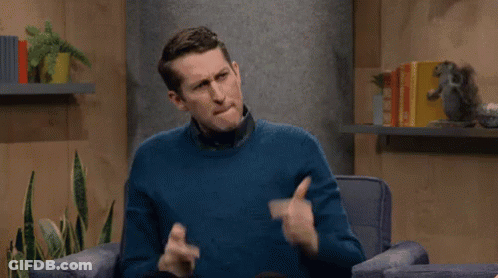 A Man In A Blue Sweater Is Sitting In A Chair Making A Gesture With His Hands . GIF