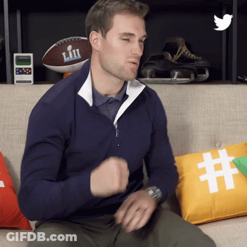 A Man In A Blue Sweater Is Sitting On A Couch With A Pillow That Says # On It GIF