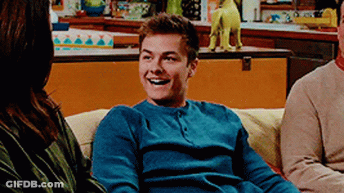 A Man In A Blue Sweater Is Smiling While Sitting On A Couch . GIF
