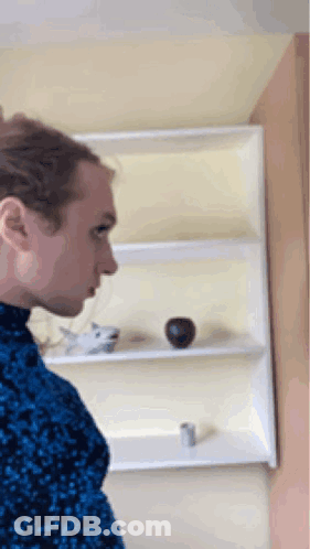 A Man In A Blue Sweater Is Standing In Front Of A Shelf With A Vase On It GIF