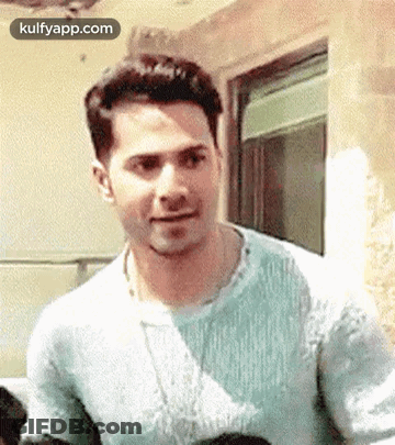 A Man In A Blue Sweater Is Standing In Front Of A Window And Making A Funny Face . GIF