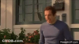 A Man In A Blue Sweater Is Standing In Front Of A Window With Shutters On It . GIF