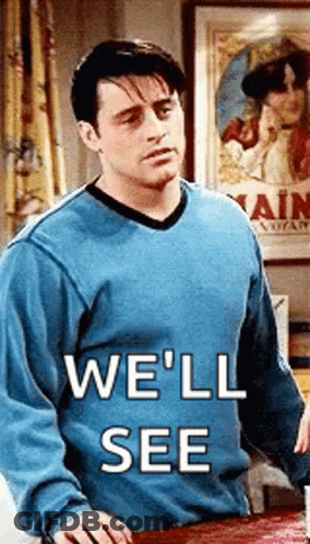 A Man In A Blue Sweater Is Standing Next To A Table With The Words `` We 'Ll See '' Written On It . GIF