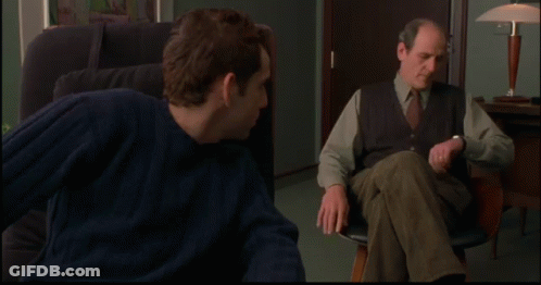 A Man In A Blue Sweater Sits In A Chair Next To Another Man In A Vest And Tie GIF