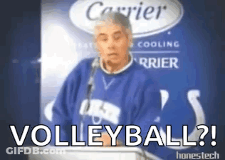 A Man In A Blue Sweater Stands At A Podium With The Words " Volleyball " Written On It GIF
