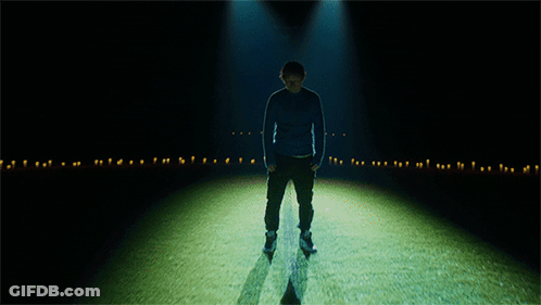 A Man In A Blue Sweater Stands In A Dark Room GIF