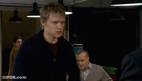 A Man In A Blue Sweater Stands In A Dark Room With Other Men GIF