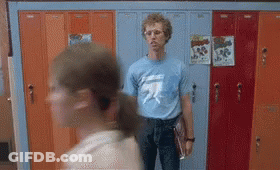 A Man In A Blue T-Shirt Is Standing In Front Of A Row Of Lockers . GIF