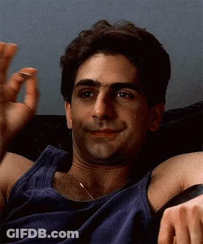 A Man In A Blue Tank Top Is Sitting On A Couch With His Arms Outstretched And Smiling . GIF