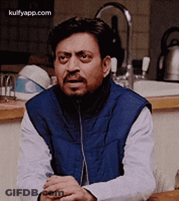 A Man In A Blue Vest Is Sitting At A Table With His Hands Folded In Front Of A Sink In A Kitchen . GIF