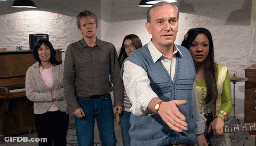 A Man In A Blue Vest Stands In Front Of A Group Of People Including A Woman Playing A Guitar GIF