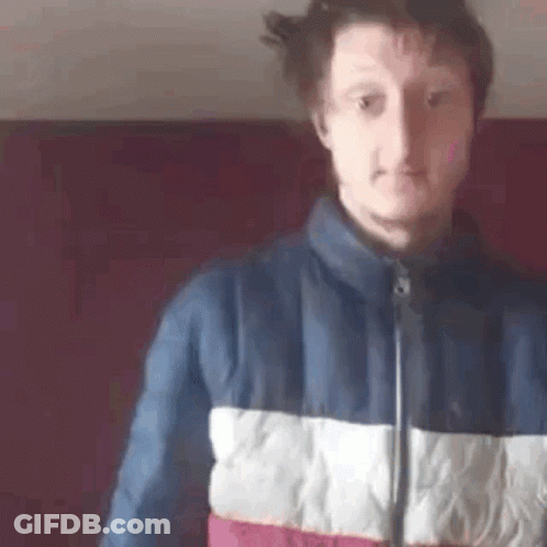 A Man In A Blue , White , And Red Jacket Is Standing In Front Of A Red Wall . GIF