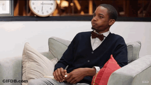 A Man In A Bow Tie Sits On A Couch In Front Of A Clock That Shows The Time As 4:20 GIF