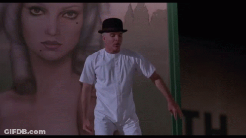 A Man In A Bowler Hat Is Standing In Front Of A Sign That Says Look Up Here GIF