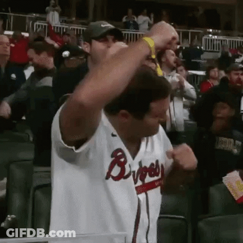 A Man In A Braves Jersey Is Standing In The Stands With His Arms In The Air . GIF