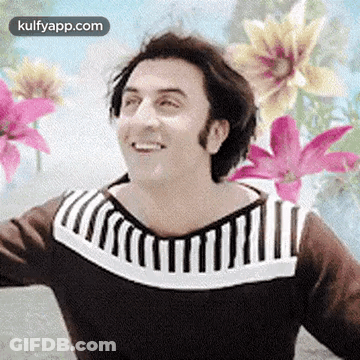 A Man In A Brown And White Striped Sweater Is Smiling In Front Of Flowers . GIF
