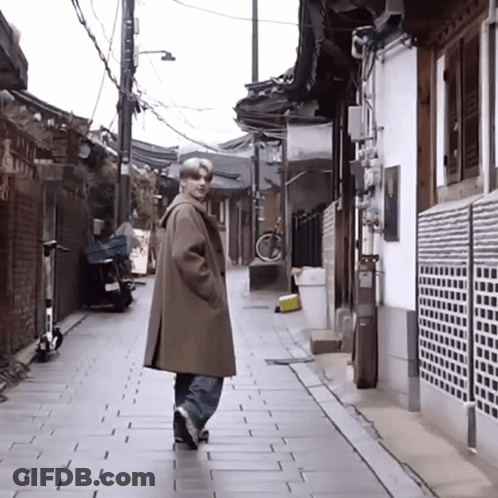 A Man In A Brown Coat Is Walking Down A Narrow Alleyway GIF