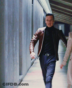 A Man In A Brown Jacket And Black Turtleneck Is Walking Down A Hallway GIF